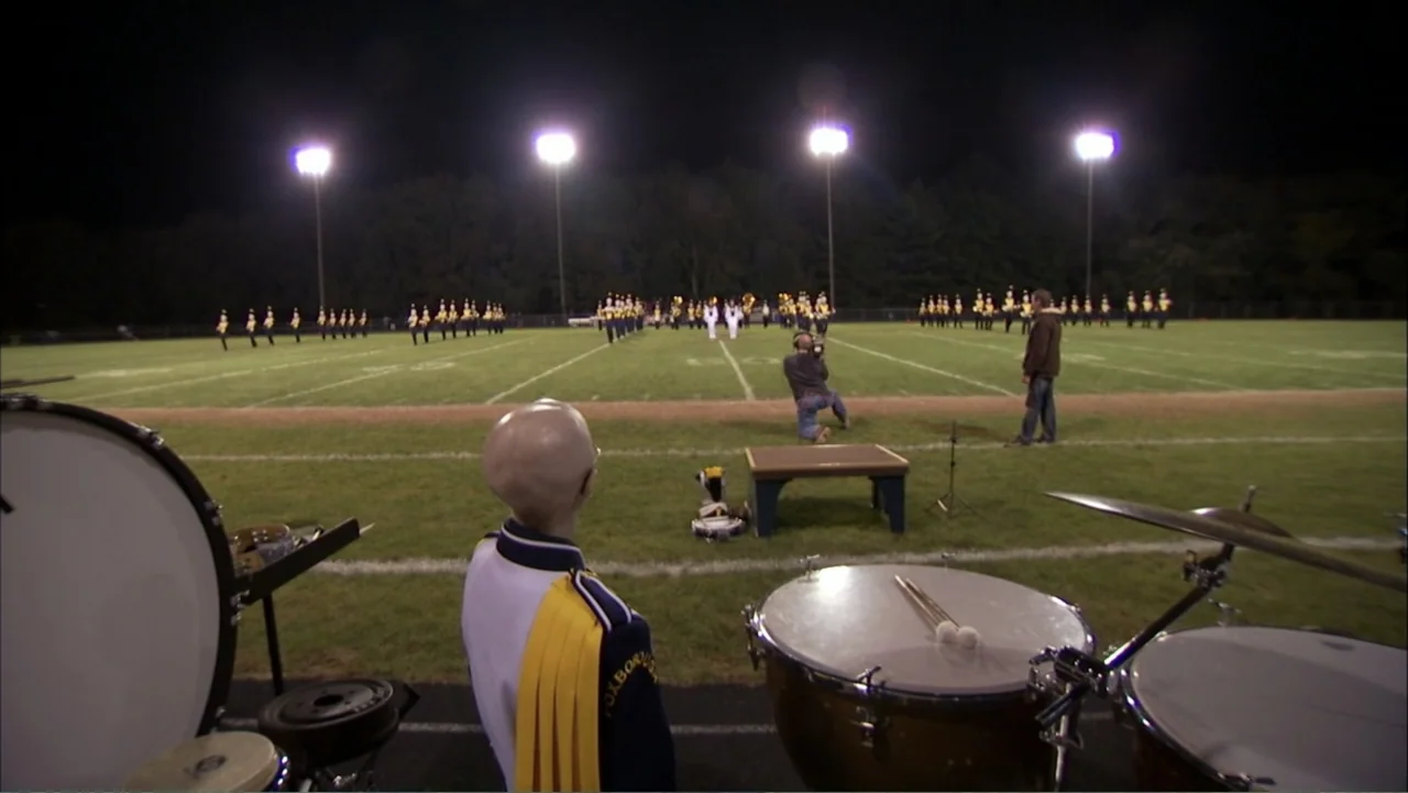 Life According to Sam (2013) movie still: medium — A night view of a grassy sports field under tall stadium lighting fixtures. A bald student in a…