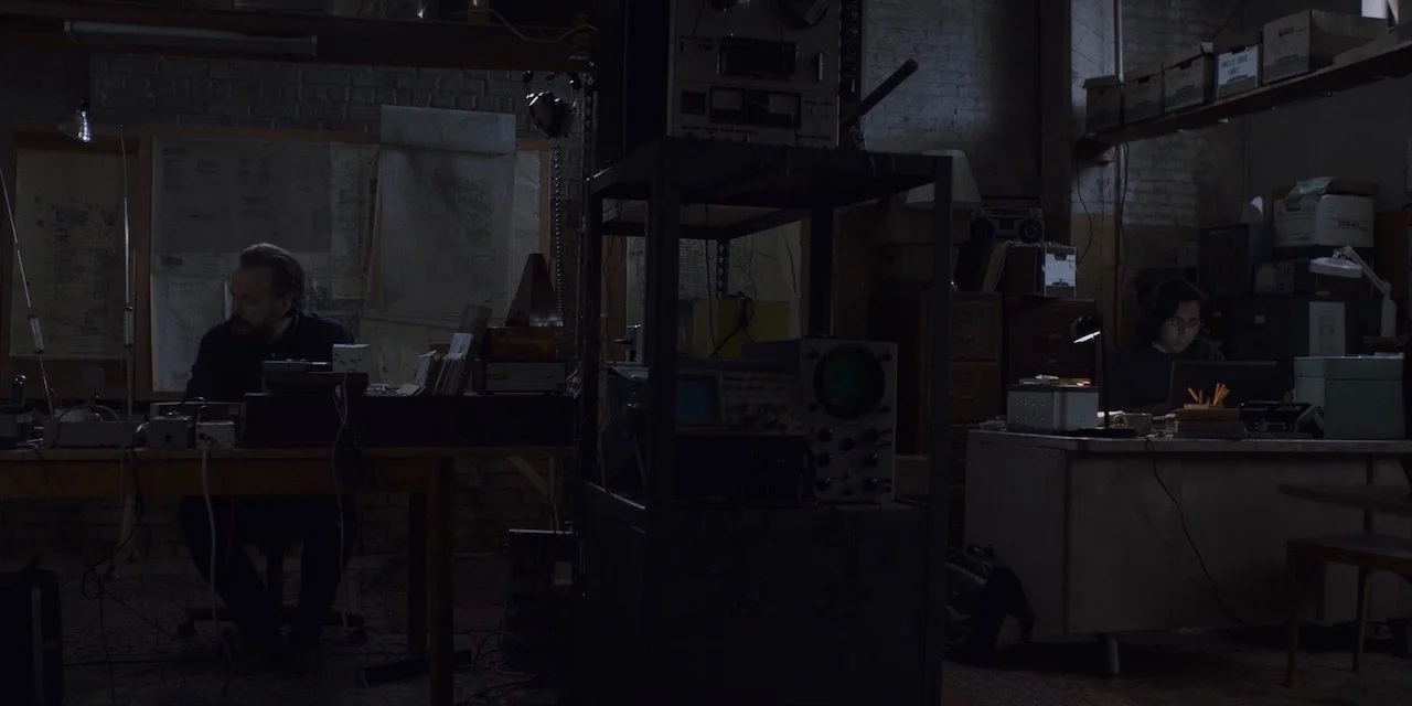 The Sound of Silence movie still: wide — A dimly lit industrial room filled with vintage electronic equipment and stacked storage boxes. Two men sit…