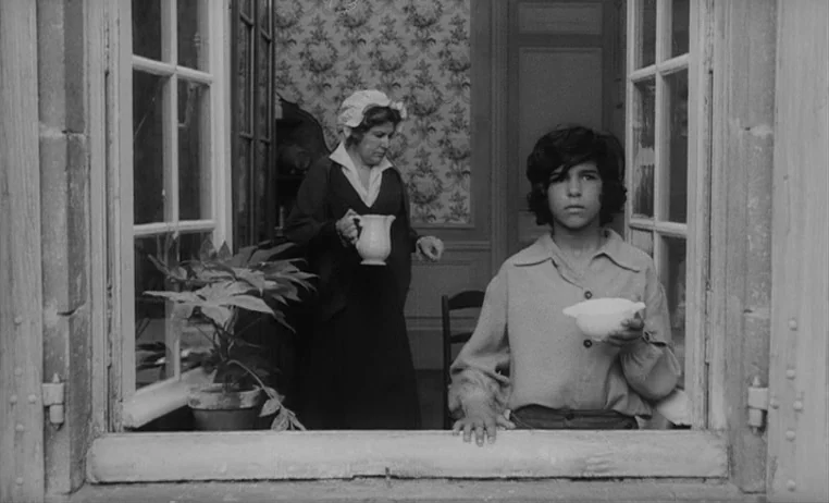 The Wild Child (1970) movie still: medium — An interior space viewed from outside through a wooden-framed window in an old house. A young boy stands…