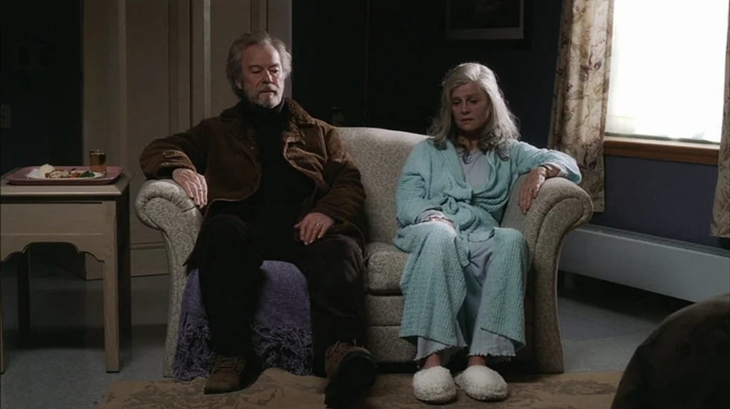Away From Her (2006) movie still: medium — A residential room features a patterned sofa placed against a dark blue wall. An elderly couple sits…