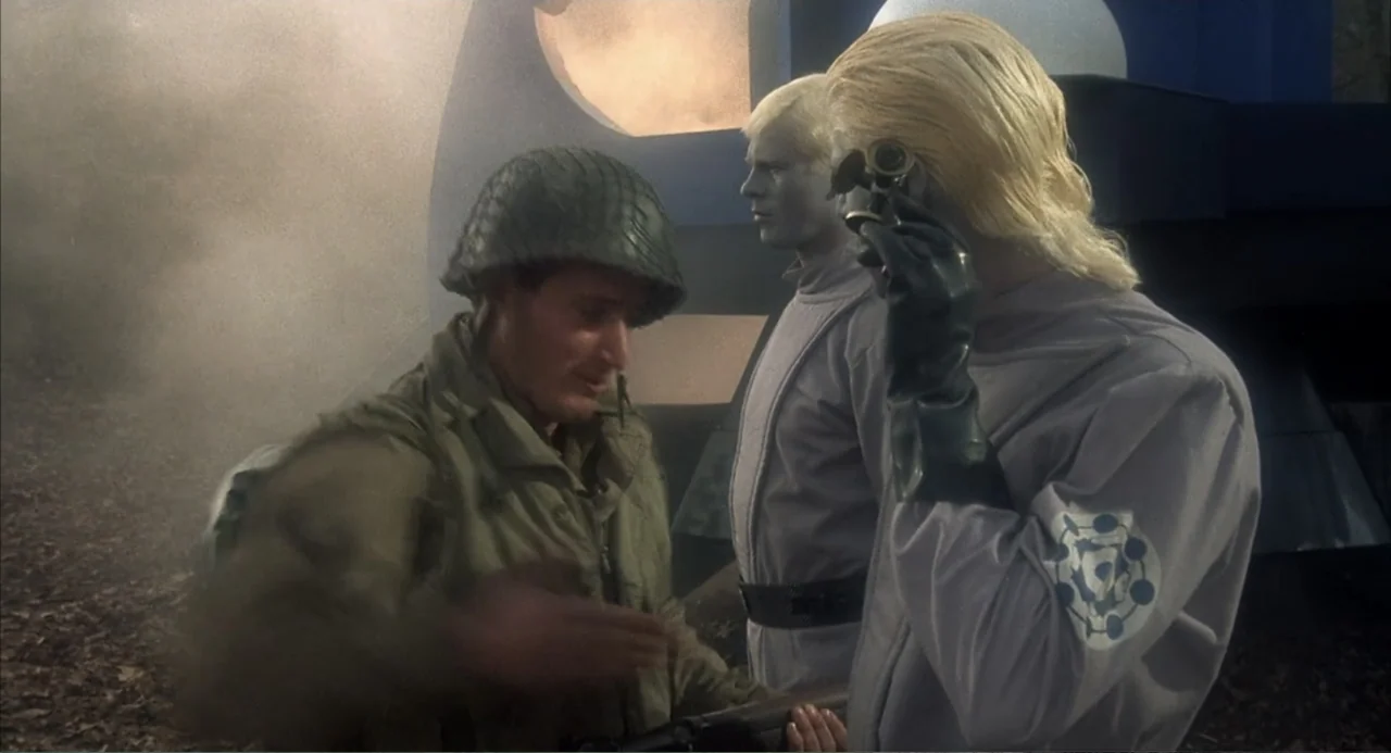 Zone Troopers (1985) movie still: medium — A wooded area with hazy air and foliage on the ground. A soldier in olive drab gear interacts with two…