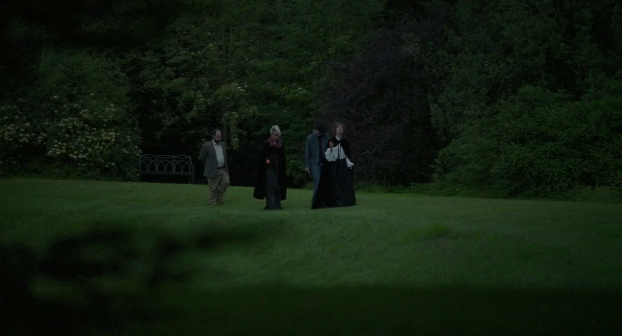 Flux Gourmet (2022) movie still: wide — A group of four people walk across a grassy expanse during the blue hour. Dense trees and a wrought iron…