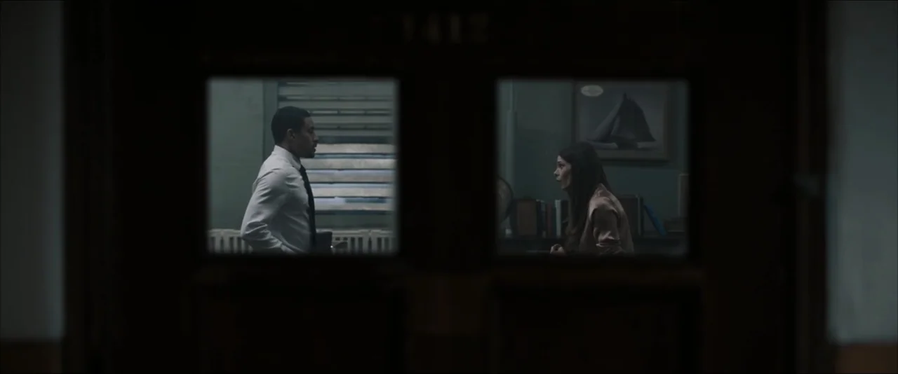 It Feeds (2025) movie still: medium — A dark, framed doorway provides a view into an office with muted green walls. A man in a white dress shirt and…