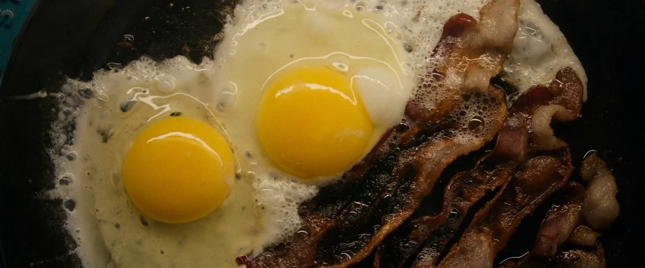 Don’t Worry Darling (2022) movie still: closeup — Two eggs and several strips of bacon cook in a dark, seasoned cast iron skillet. The edges of the…