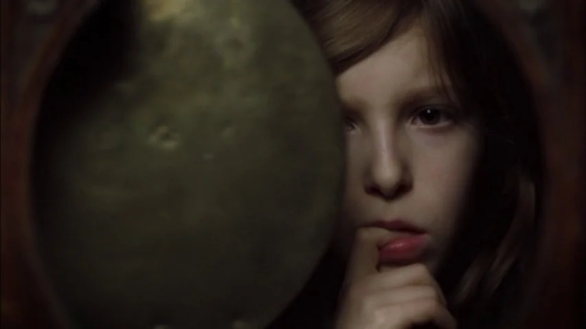 The Sleeping Beauty (2010) movie still: closeup — A dark space features a young girl partially hidden behind a circular wooden frame. She holds one…