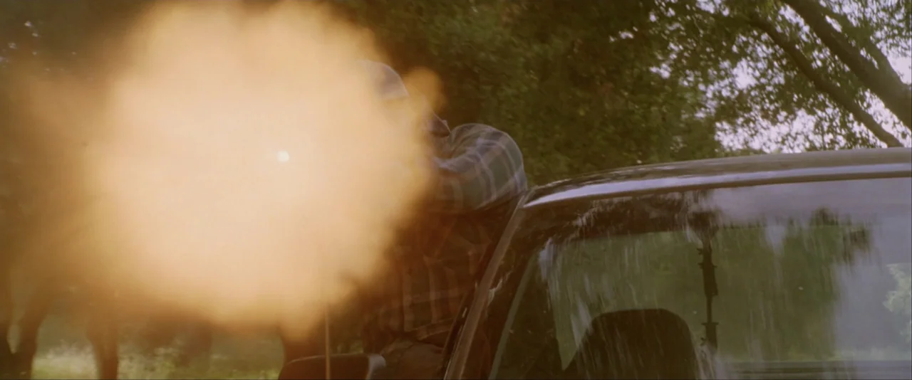 The Dukes of Hazzard (2005) movie still: medium — A person in a plaid shirt fires a rifle from a car window outdoors. The bright muzzle flash…