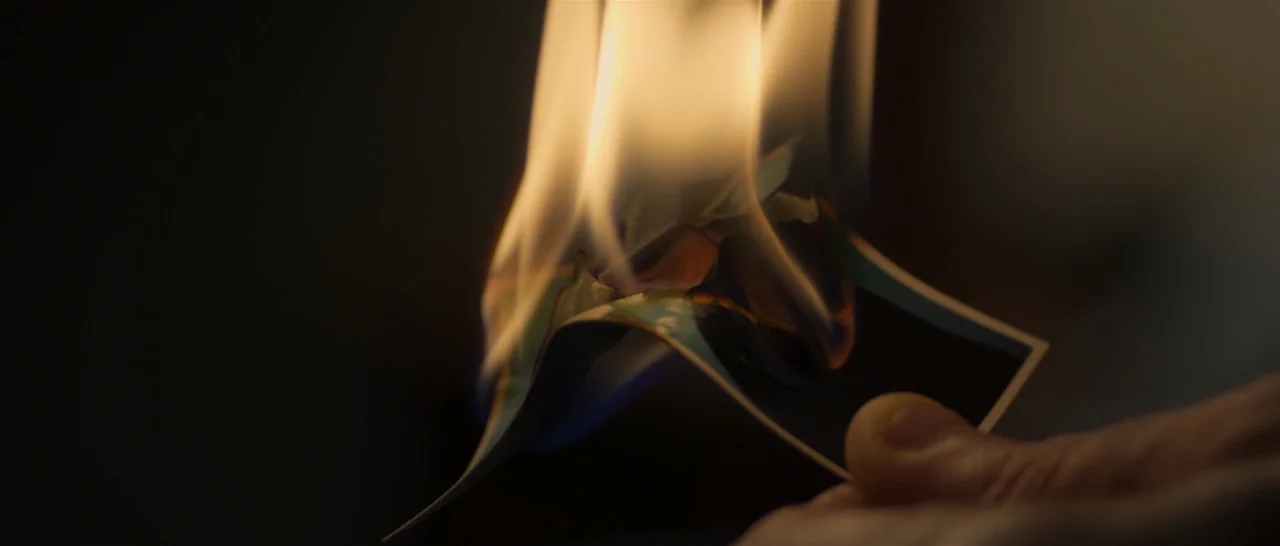 You Were Never Really Here (2017) movie still: closeup — A close view shows a small square photograph being consumed by bright yellow flames. The…