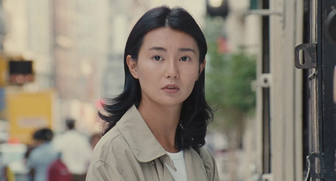 Comrades: Almost A Love Story (1996) movie still: closeup — An outdoor city sidewalk with blurred urban architecture and indistinct pedestrians in…