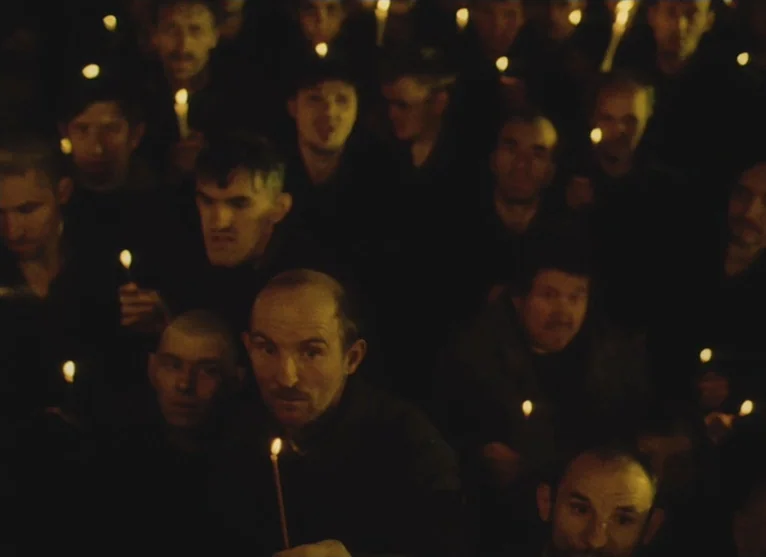 A Visitor to a Museum (1989) movie still: medium — A dense crowd of men packed closely together in a dimly lit dark space. Each individual holds a…
