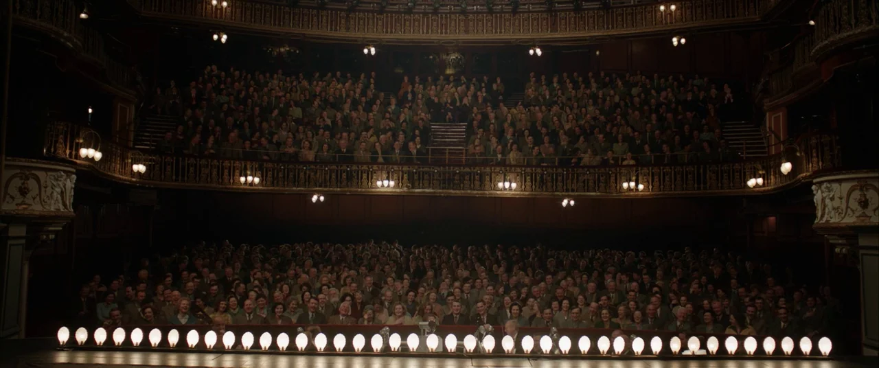 Stan & Ollie (2018) movie still: wide — A classic ornate auditorium filled with a dense crowd of audience members. Rows of warm incandescent…