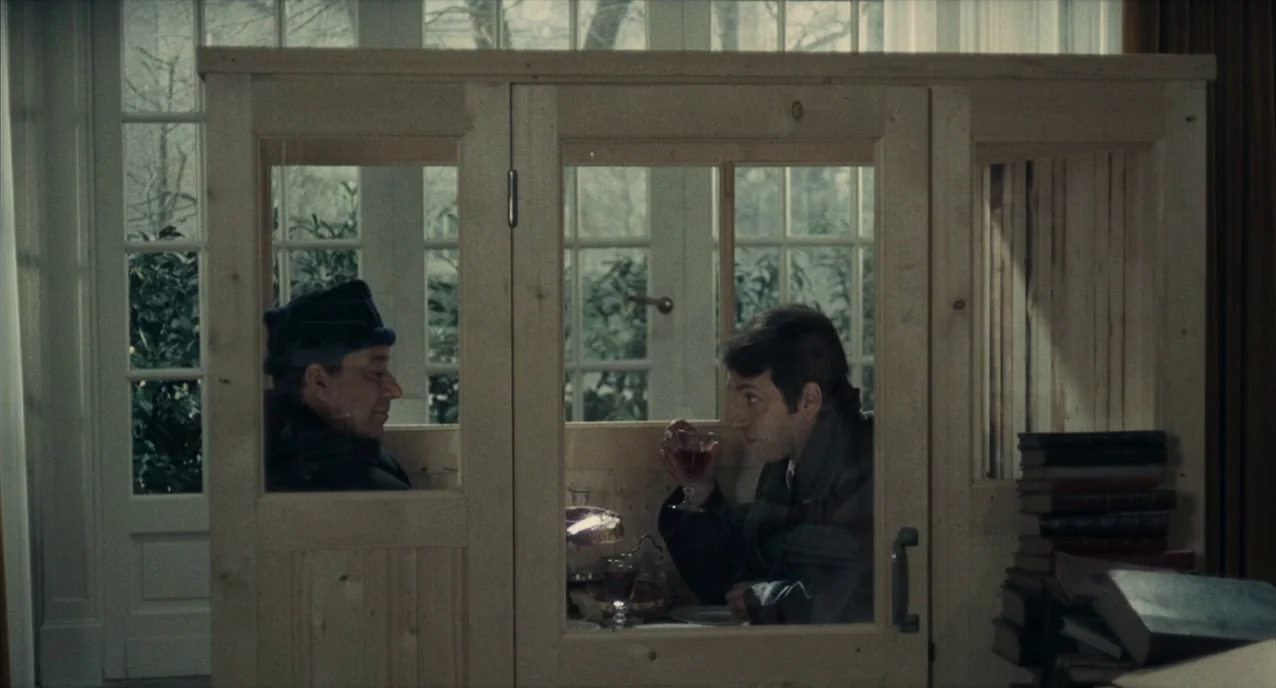The Army of Shadows (1969) movie still: medium — A light wood, partitioned booth with glass panels stands in a bright room. Two men sit inside the…