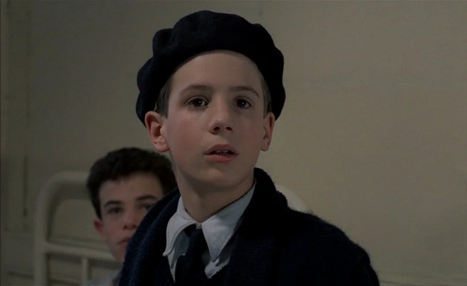 Au Revoir Les Enfants (1987) movie still: closeup — A plain room with light colored walls and metal bed frames in the background. A young boy in a…