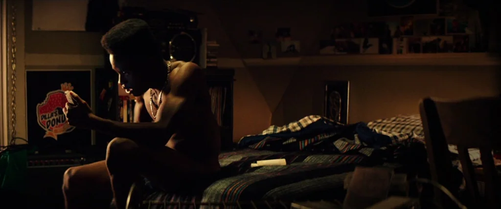 Dope (2015) movie still: medium — A dimly lit room with 1980s period items like a boombox and posters on walls. A shirtless man sits on a plaid bed…