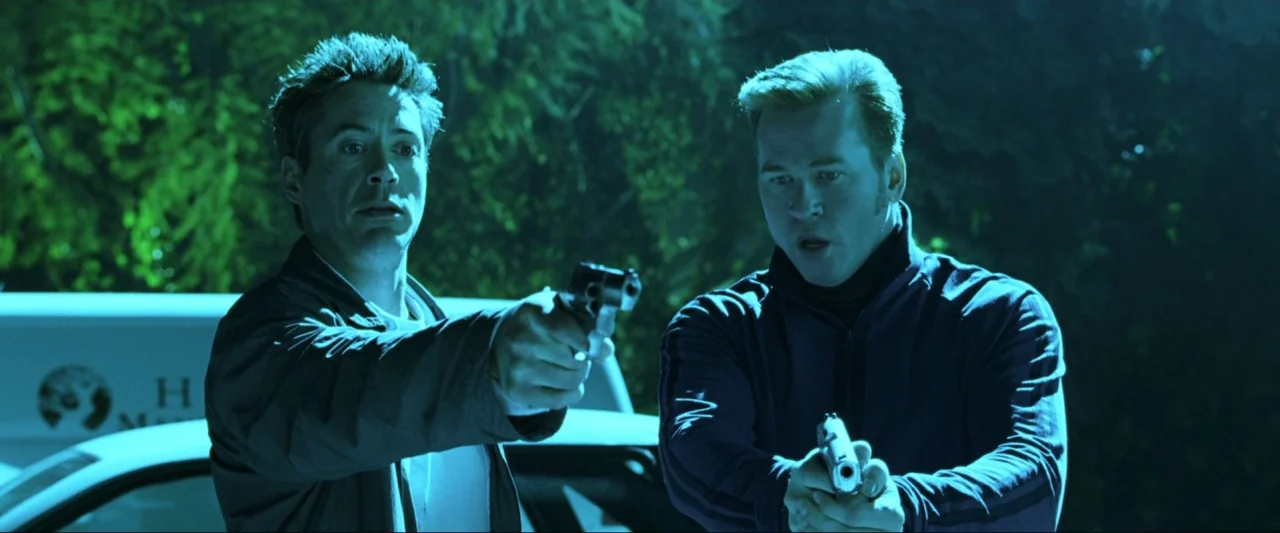 Kiss Kiss Bang Bang (2005) movie still: medium — Two men stand at night in an outdoor parking area near a white vehicle. They hold pistols with both…