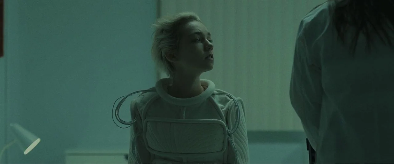 Come True (2020) movie still: medium — A clinical examination room features green walls and a desk lamp in the corner. A woman with short blonde…