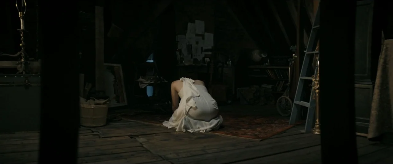 It Feeds (2025) movie still: medium — A cluttered, shadowy attic space features wooden rafters, storage boxes, and a ladder. A woman in a white…