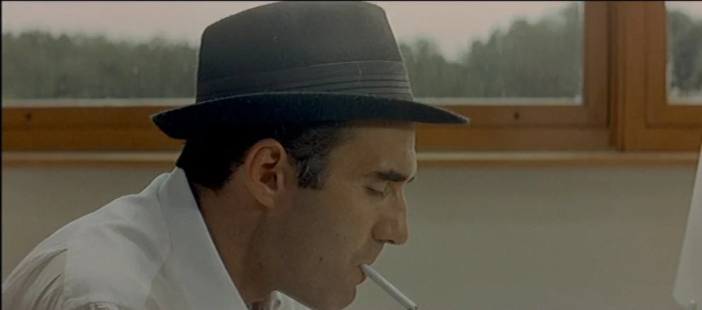 Contempt (Le Mépris) (1963) movie still: closeup — A man sits indoors near a large window looking out at a treeline. He wears a fedora and holds a…