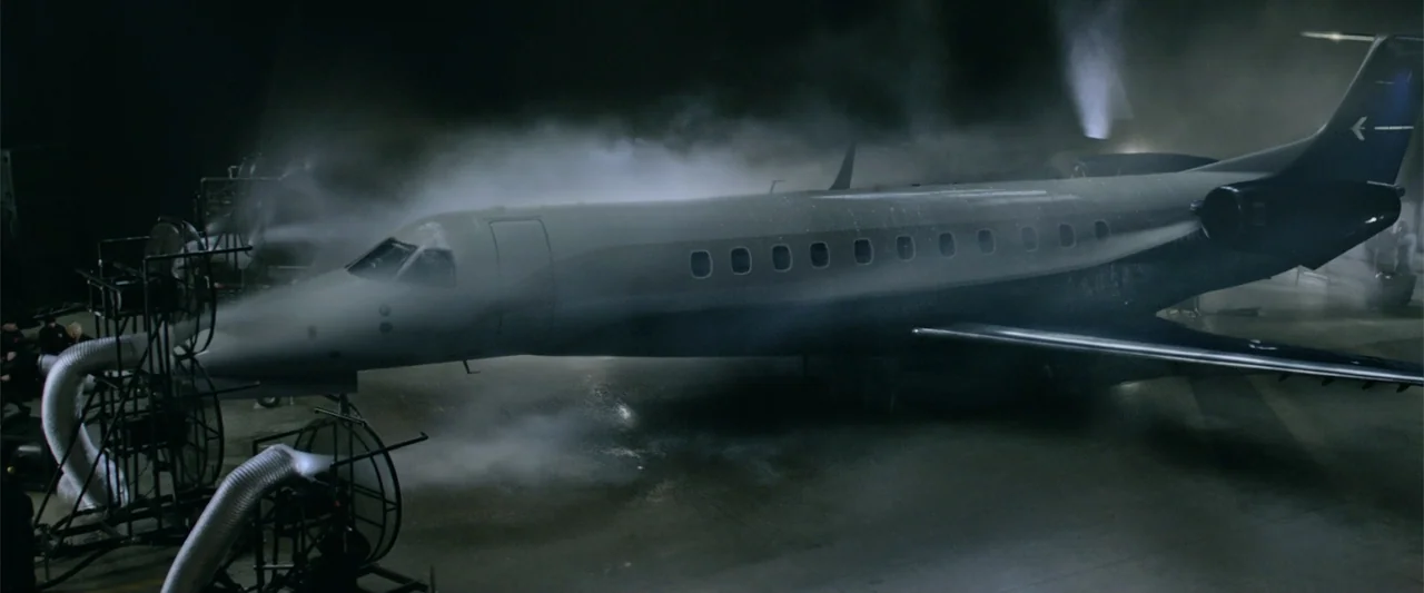 Now You See Me 2 (2016) movie still: wide — A private jet sits inside a dark, concrete-floored hangar with industrial machinery nearby. Thick fog or…