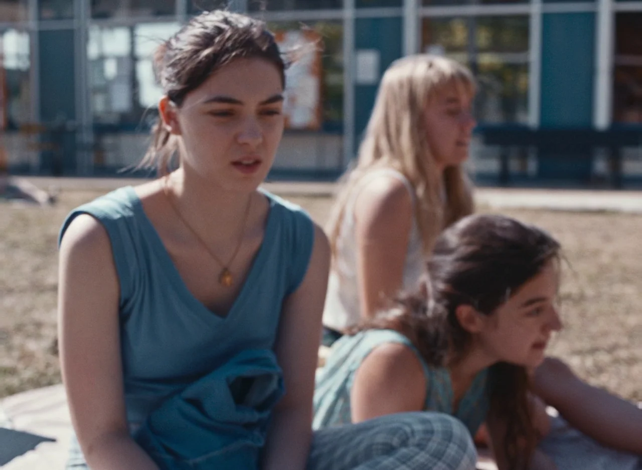 Happening (2021) movie still: medium — A school courtyard with large glass windows visible in the background structure. Three teenage girls sit…