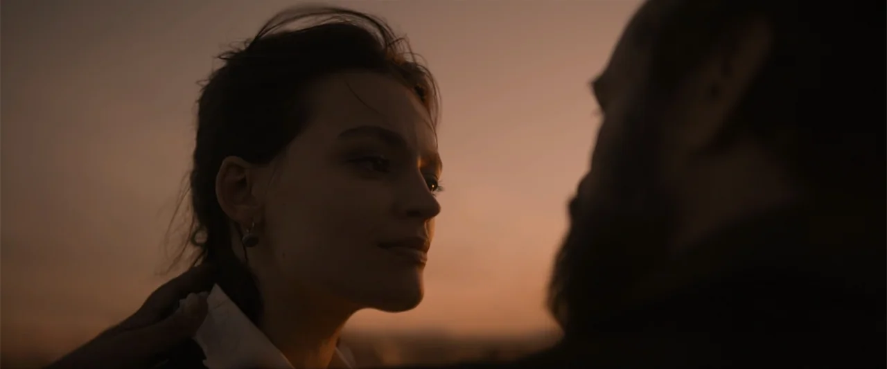 Eiffel (2021) movie still: closeup — An outdoor terrace during golden hour with a clear sky in the background. A man reaches out to touch the neck…