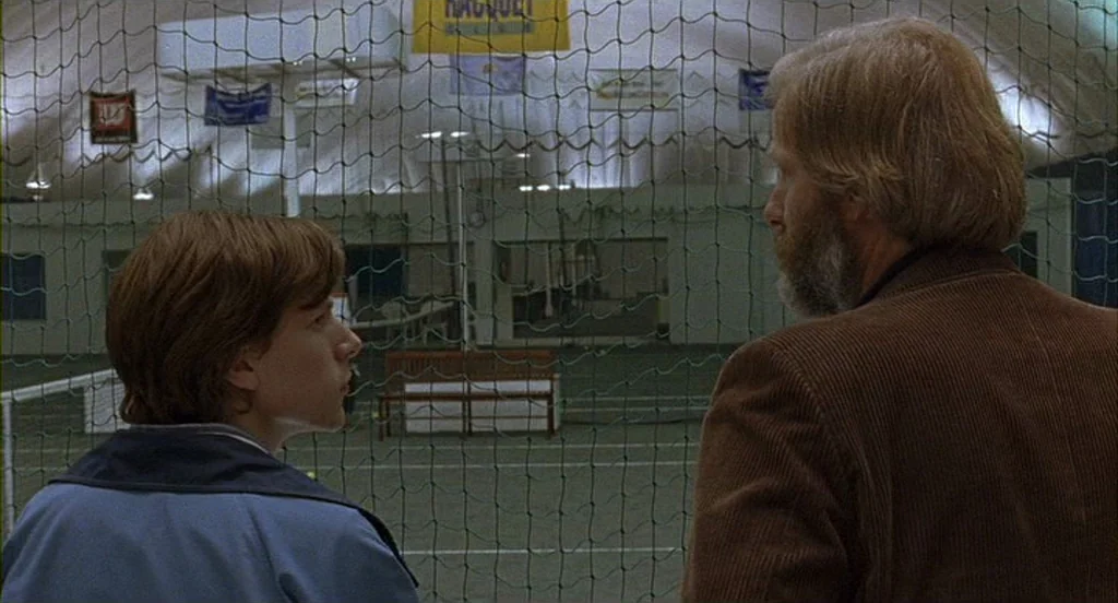 The Squid and the Whale (2005) movie still: medium — An indoor tennis facility with a protective green nylon net separating the subjects. A man and…