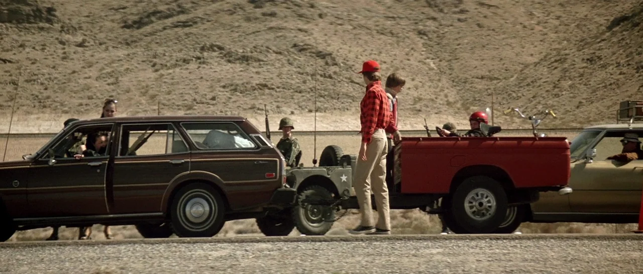 Starman (1984) movie still: wide — A group of people gathers around a brown station wagon and a red pickup truck. They stand on a dry, rocky desert…