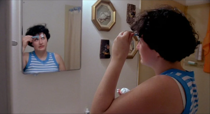 Gummo (1997) movie still: medium — A plain wall in a bathroom decorated with framed decorative pictures and small ornaments. A young woman uses a…