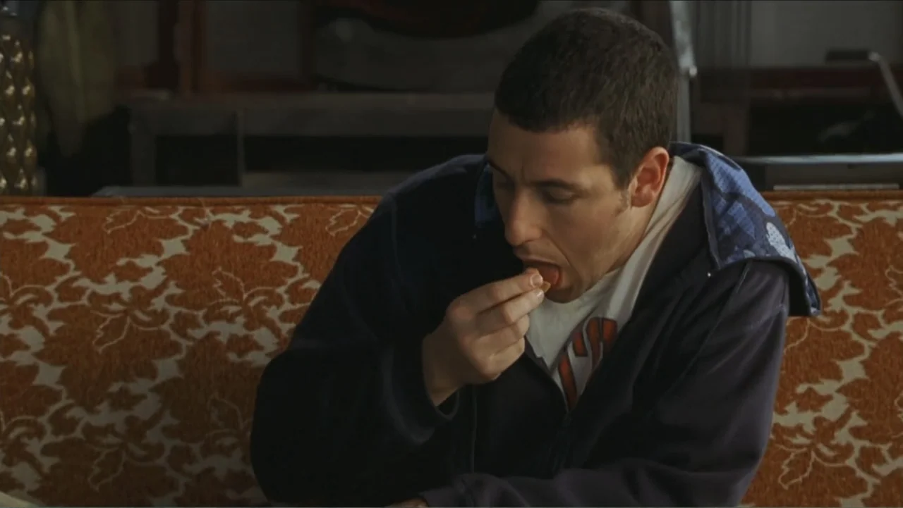 Big Daddy (1999) movie still: medium — A man sits on a patterned orange sofa in a room. The man eats a small piece of food from his fingers.
