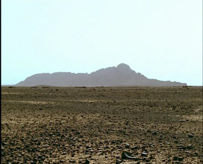 Fata Morgana (1971) movie still: wide — A vast, arid desert plain covered in loose rocks and sparse scrub vegetation. A large, dark rocky mountain…