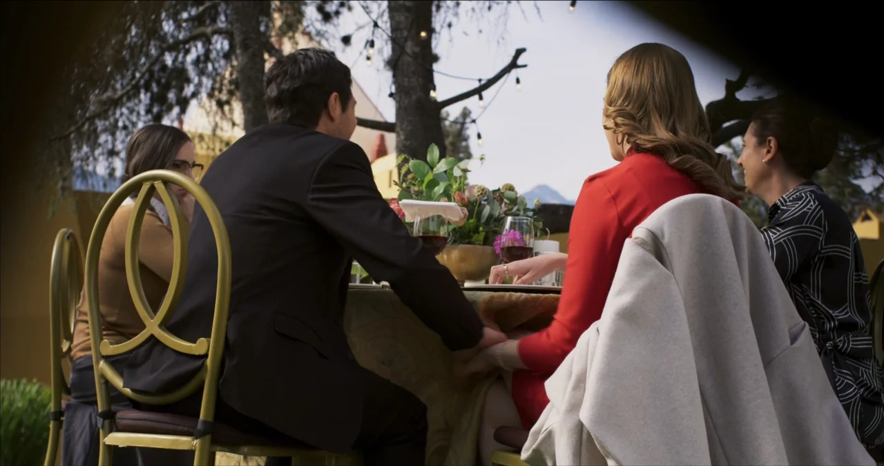 Bendita Suegra (2023) movie still: medium — An outdoor dining area features a table set with wine glasses and floral centerpieces. Four adults sit…