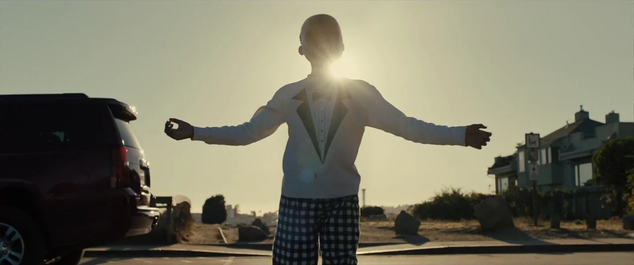 Us (2019) movie still: fullbody — A sunny parking lot borders a coastal road with houses in the background. A child stands centered with arms wide…
