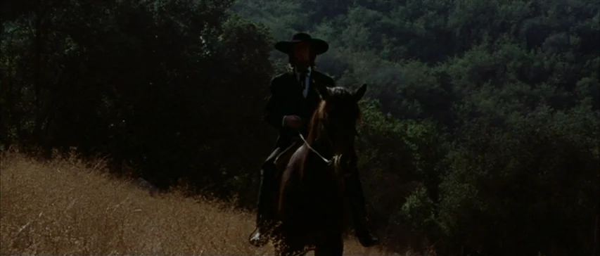 Messiah of Evil (1973) movie still: fullbody — A man rides a dark horse along a dry grassy hillside during the day. Dense green trees fill the…