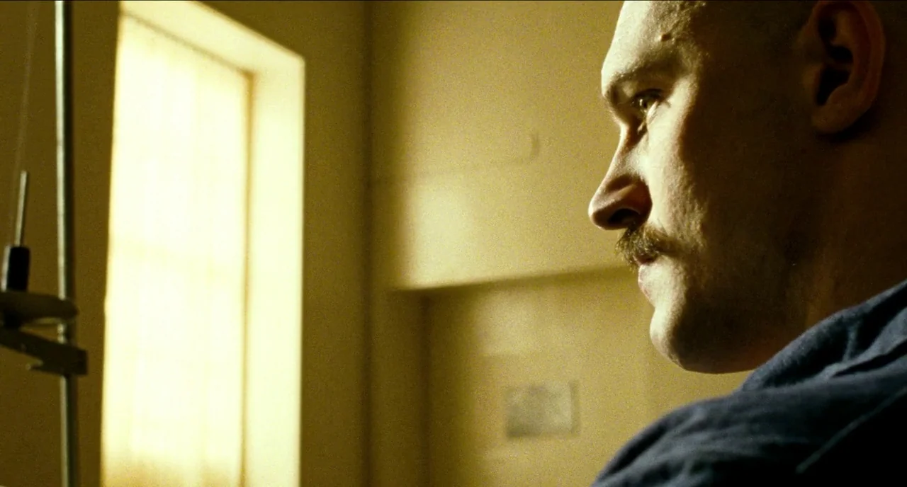 Bronson (2008) movie still: closeup — A dimly lit room features a bright window that casts a yellow glow. A man with a mustache looks off-screen…