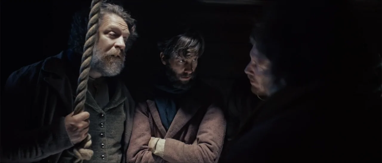 Out of Liberty (2019) movie still: medium — A dark, cramped interior with wooden walls and low overhead lighting. Three men stand close together in…