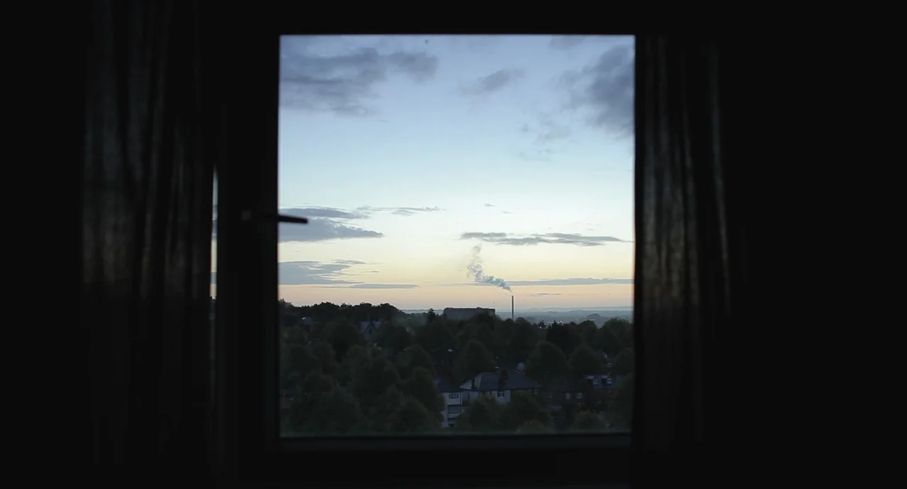 Weekend (2011) movie still: establishing — A dark, silhouetted window frame looks out over a suburban landscape at twilight. A distant chimney emits…