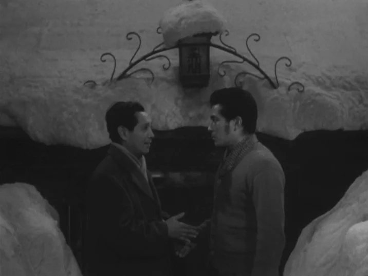 The Idiot (1951) movie still: medium — An outdoor structure covered in thick snow features a prominent central iron lantern fixture. Two men face…