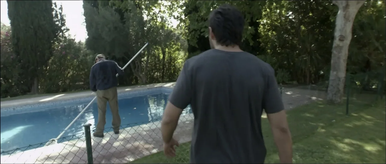 Otro verano (2013) movie still: medium — A sunny backyard features a swimming pool surrounded by mature trees and a chain-link fence. One man…