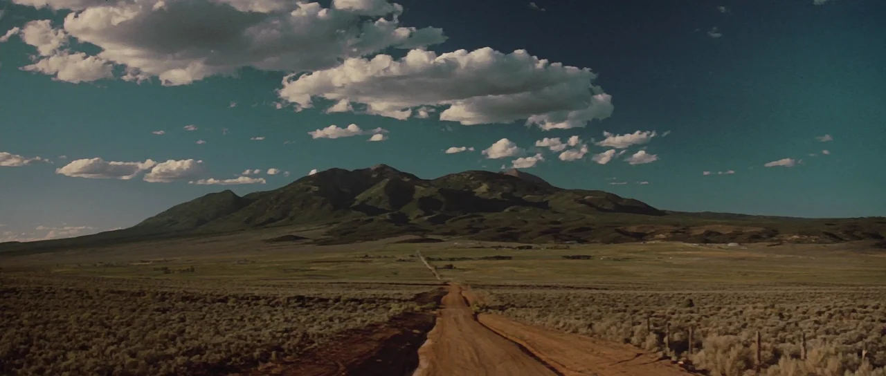 Thelma & Louise (1991) movie still: establishing — A wide, unpaved dirt road cuts through a vast, grassy plain under a large mountain. Wispy white…