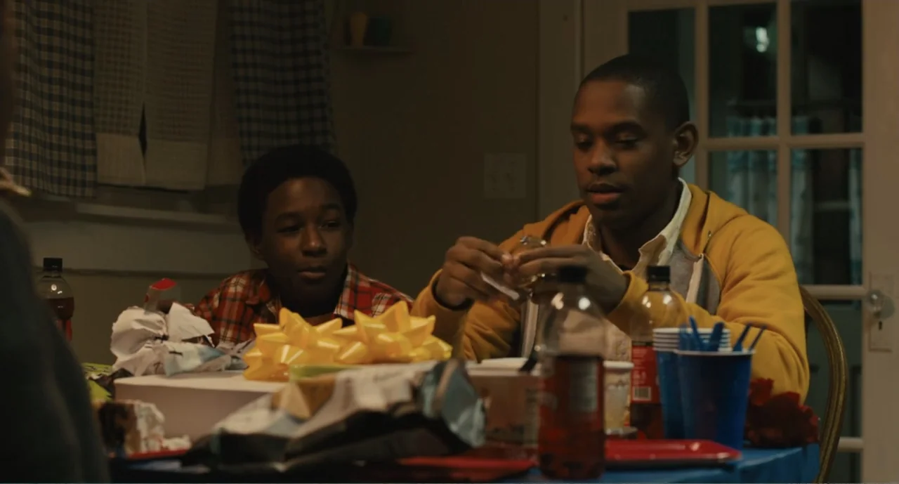 Lila & Eve (2015) movie still: medium — A domestic dining area contains a table covered with wrapping paper and gifts. Two young men sit at the…