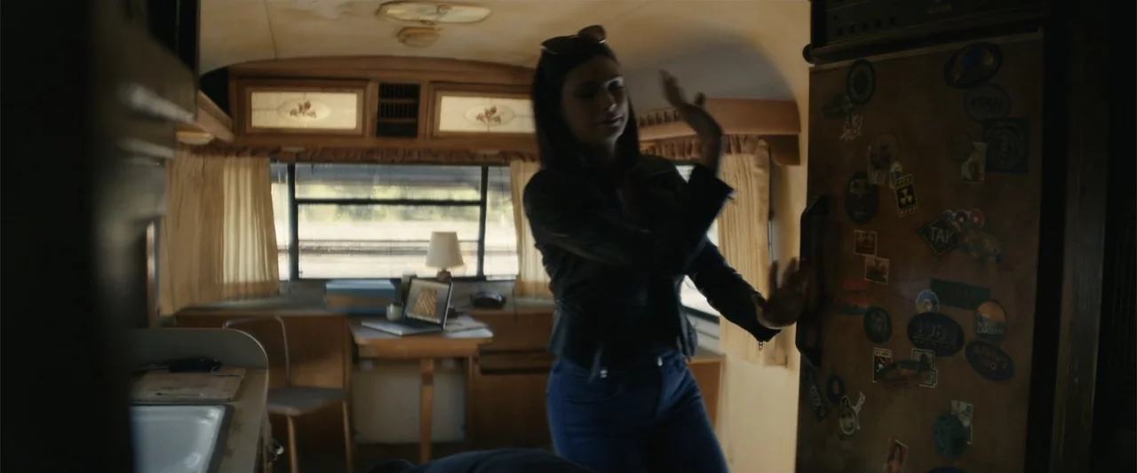 Last Looks (2022) movie still: medium — An interior of a compact camper trailer contains a cluttered refrigerator and a desk. A young woman wearing…