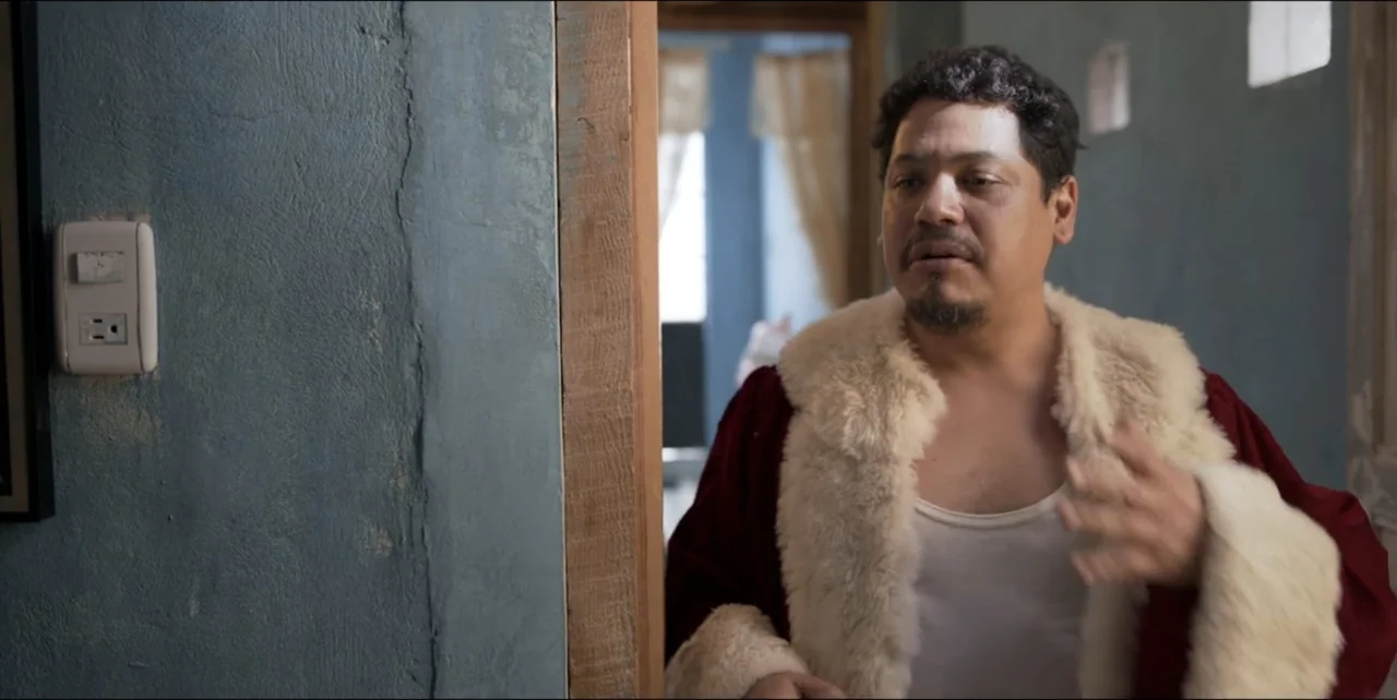 El sabor de la Navidad (2023) movie still: medium — A residential interior corridor features textured blue walls and a wooden door frame. A man in a…