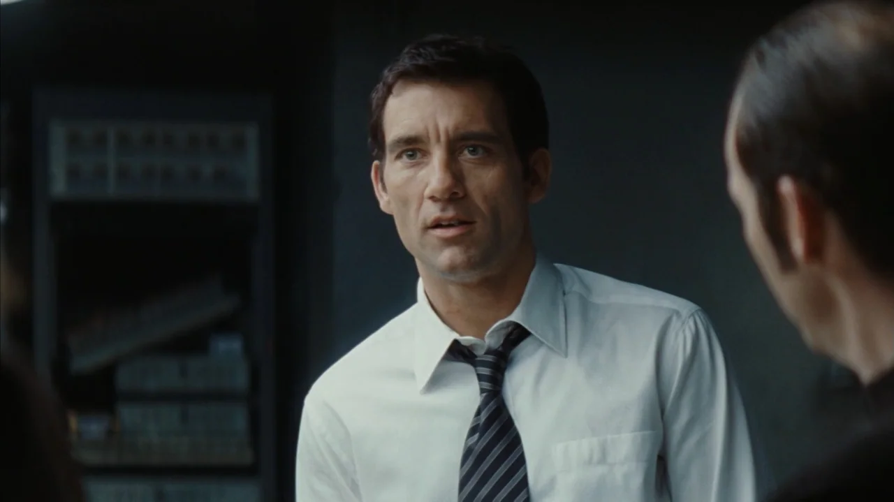 Duplicity (2009) movie still: medium — A dark office environment features a man in professional attire engaged in a conversation. The man stands…