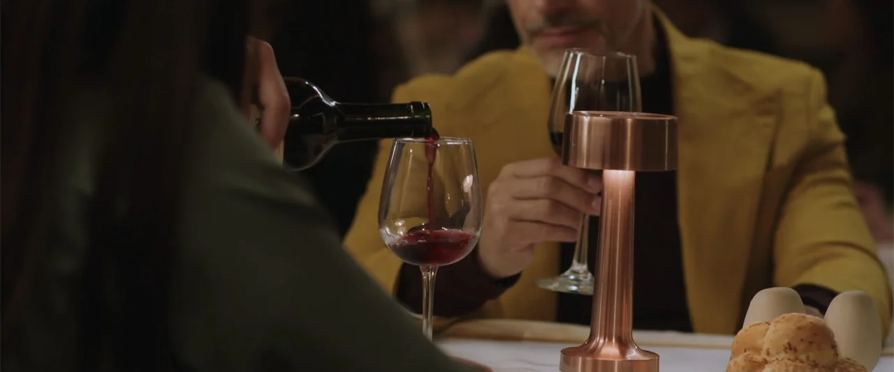 Winner Takes the Cake (2025) movie still: medium — A dimly lit table at a restaurant features a modern copper-colored metallic desk lamp. One person…