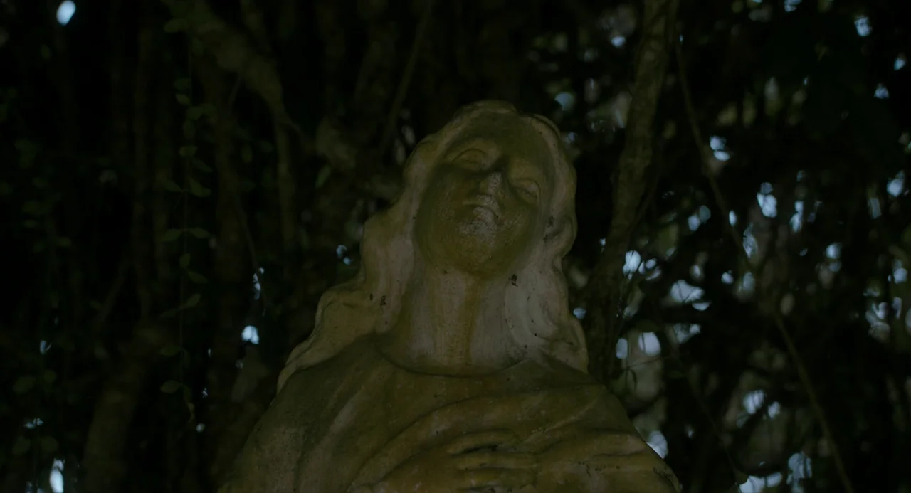 In My Mother’s Skin (2023) movie still: closeup — An weathered stone statue of a woman rests amidst dark, dense foliage. The figure is captured from…