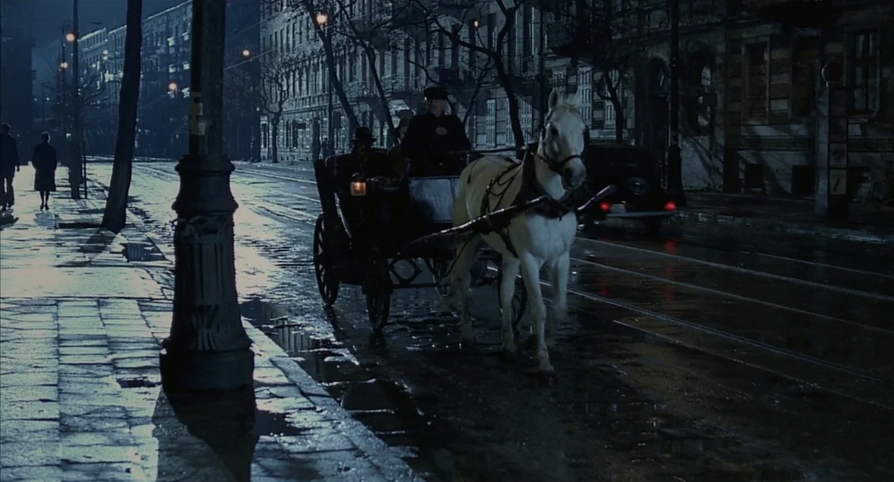 The Pianist (2002) movie still: wide — A wet cobblestone city street at night is illuminated by dim, distant streetlamp light. A white horse pulls a…