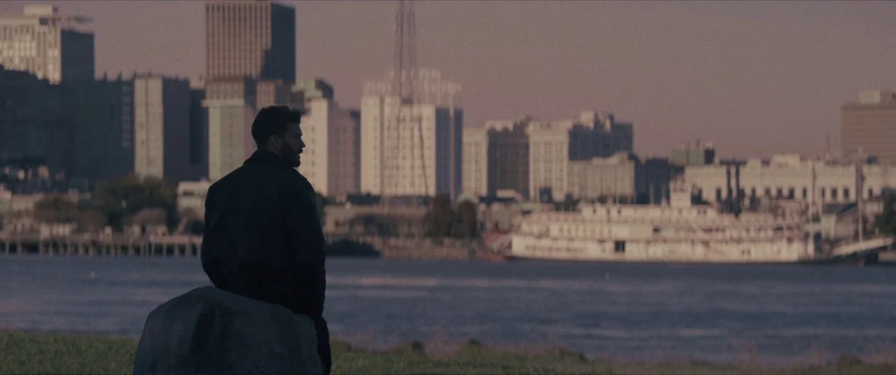 Synchronic (2019) movie still: medium — A grassy riverbank overlooks a wide river with a cityscape in the distance. A man sits on a large rock…