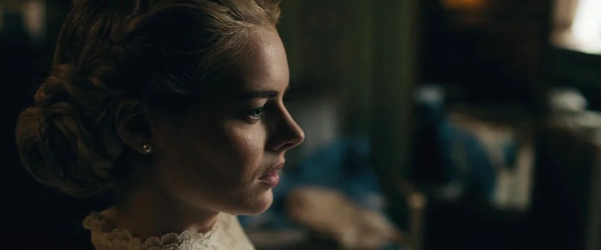 Ready or Not (2019) movie still: closeup — An interior space with dimly lit background elements obscured by a shallow depth of field. A woman in a…