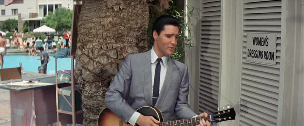 Viva Las Vegas (1964) movie still: medium — A sunny pool deck features a tall palm tree next to a dressing room door. A man in a suit stands by the…