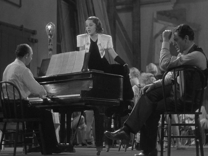 42nd Street (1933) movie still: medium — A rehearsal room features a grand piano and wooden chairs in a sparse space. A woman performs a song while…