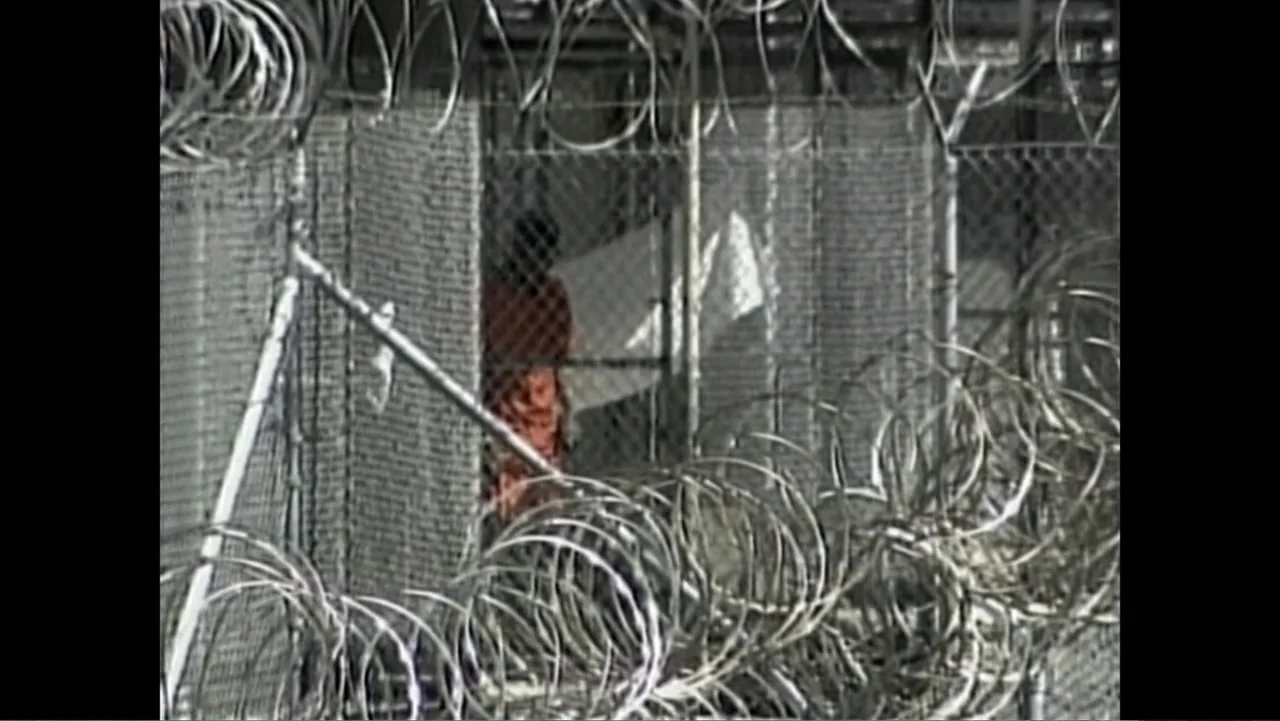 The Forever Prisoner (2021) movie still: wide — A high-security area features multiple layers of chain-link and circular razor wire. A single person…